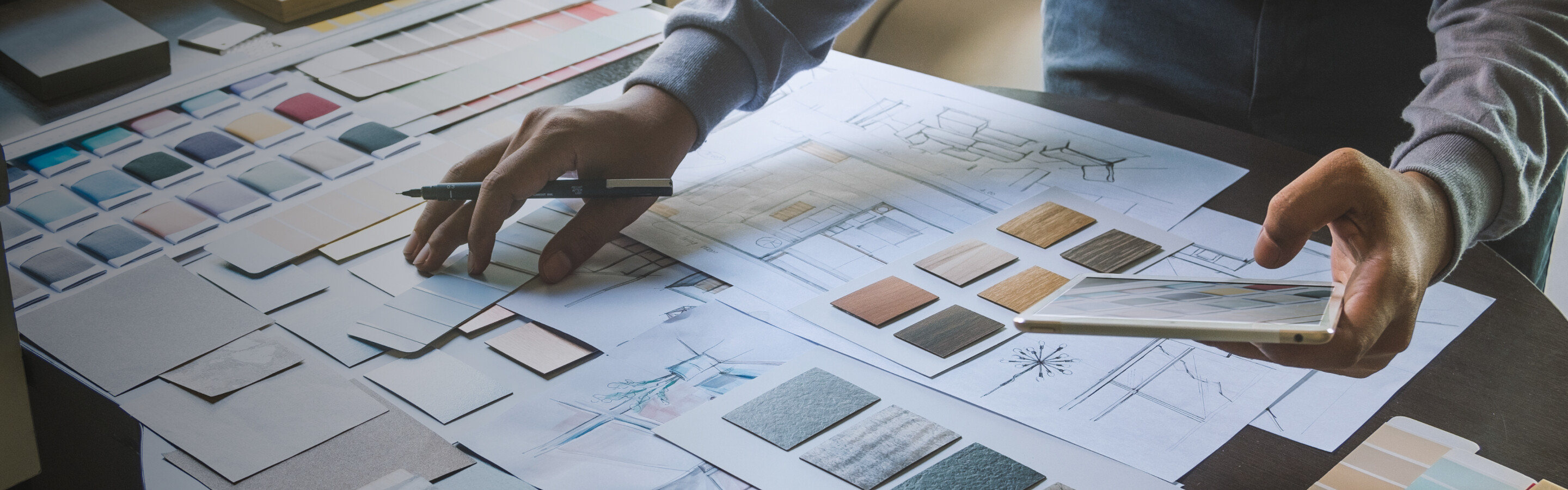 A shot of a table with interior design drawings on it and someones arm holding a pencil